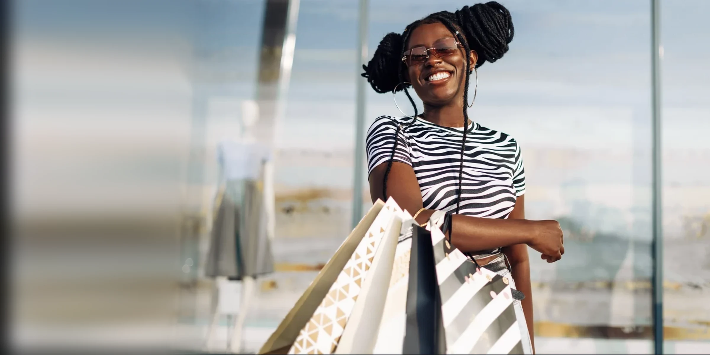 A smiling shopper in a zebra-print top and sunglasses holding paper shopping bags in front of a shop window.